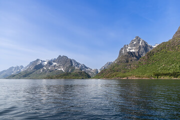 Lofoten's towering mountains, with their snowy crevices, rise majestically above the tranquil fjord, embodying the serene spirit of Norway's natural grandeur