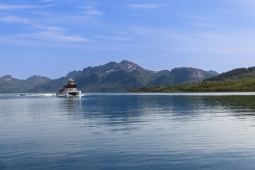 A catamaran glides through the glassy waters of the Lofoten fjords, nestled among the verdant,...