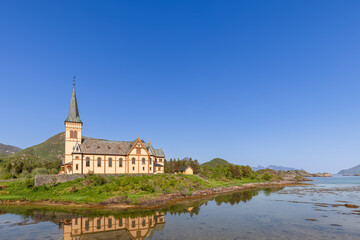 Vagan Kirke, nestled in Lofoten's stunning landscape, reflects its traditional architecture in the tranquil waters on a bright Norwegian day