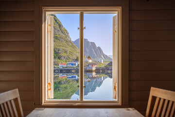 Inside a rorbu, looking through an open window, the calm Norwegian fjord mirrors quaint houses and a rugged cliff under a clear sky