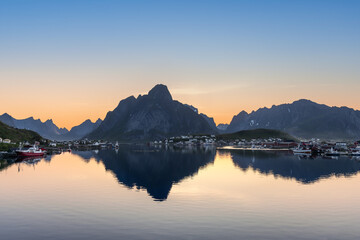 Dawn breaks over a tranquil Norwegian fjord in Lofoten, with the serene waters reflecting the silhouette of rugged peaks and a sleepy fishing village awakening to the soft hues of sunrise
