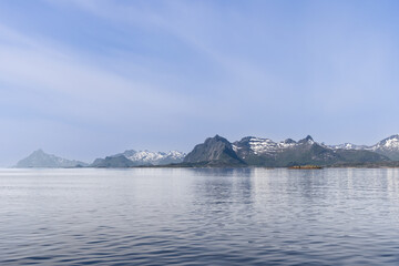 The Lofoten Islands bask in the quiet of summer, with gentle fjord waters reflecting the majestic, snow-flecked mountains under a soft, diffuse light. Norway