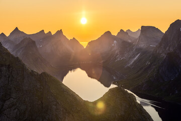Sunset bathes the jagged peaks of Reinebringen in Lofoten Islands, Norway with golden light, the midnight sun casting a serene glow over the calm waters below