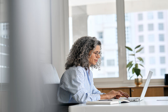Busy mature senior business woman working in office using laptop. Middle aged old professional lady executive manager looking at computer digital technology sitting at desk. Authentic candid shot.