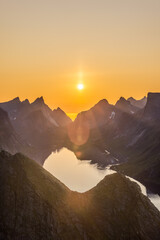 The vertical frame captures the midnight sun casting a warm glow over Reinebringen's sharp peaks in Lofoten, Norway, with the sun's reflection shimmering in the serene fjord