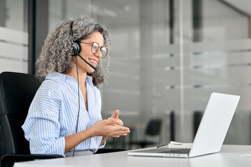 Smiling middle aged female support service employee talking to customer sitting at desk. Happy senior old woman professional call contact center agent wearing headset hybrid working in business office - obrazy, fototapety, plakaty