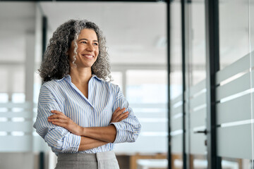 Smiling happy confident old mature professional business woman corporate leader, senior middle aged female executive, lady bank manager standing in office hallway arms crossed looking away, portrait.