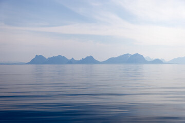 A serene view of the Lofoten Islands' silhouette under a pastel azure sky, reflected gently on the...