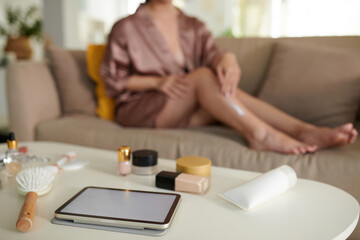 Obraz na płótnie Canvas Tablet and cosmetics on coffee table of young woman applying body lotion in background