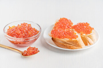 Sliced loaf of bread or canape with butter and salty salmon red caviar or organic fish roe served on plate on white wooden table with spoon and glass bowl prepared for healthy vitamin breakfast 