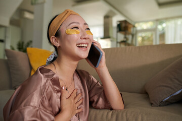 Young woman with under eye patches laughing when talking on phone with friend