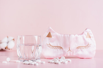 Sleeping pills, a glass of water and a sleep mask on a pink background