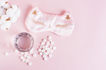 Sleeping pills, a glass of water and a sleep mask on a pink background top view