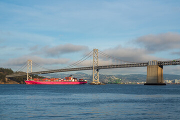 a large red container ship making its way under the Oakland Bay Bridge in daylight on a partly cloudy day.