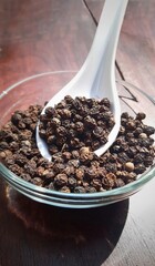 Black pepper in glass bowl. Black pepper in glass bowl and in a spoon in wooden background. Spoon and black peppercorn composition over a wooden background. Spice black pepper.