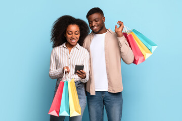 Black couple with shopping bags using smartphone, blue background