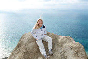 A young girl sits on a high mountain above the sea on a sunny day. Smiling blonde in a brown jacket. Active lifestyle and recreation. - Powered by Adobe