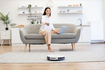 Beautiful lady switching on robot vacuum cleaner via remote control while sitting on sofa in...