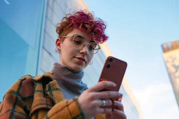 Trend-setting non-binary person with pink curls engaged with their phone against an urban backdrop.