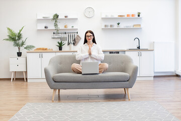 Brunette female freelancer sitting on comfy couch in namaste pose with reducing fatigue using yoga...