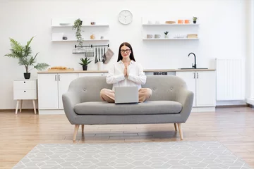 Poster Im Rahmen Brunette female freelancer sitting on comfy couch in namaste pose with reducing fatigue using yoga practice. Caucasian woman meditating with closed eyes while working on laptop at living room. © sofiko14