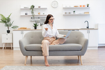 Positive brunette young female in casual attire gesturing with hands while sitting on couch with...