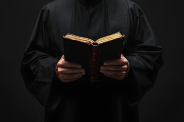 Catholic priest in black cassock robe holding the Holy Bible, isolated on a dark black background. - obrazy, fototapety, plakaty