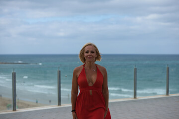 Beautiful blonde adult woman in an elegant red dress walks along the viewpoint above the beach. In the background the Atlantic Ocean in Cadiz, Spain. Travel and holiday concept.