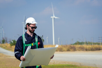 Handsome caucasian engineer, technician man wearing safety uniform workwear standing, holding and look at drawing paper or blueprint in front of windmill or turbines field with blue sky background.