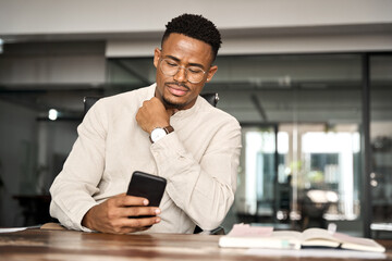 Busy professional African American business man sitting in office holding mobile cellphone. Young businessman entrepreneur using smartphone looking at cell phone technology device at work.