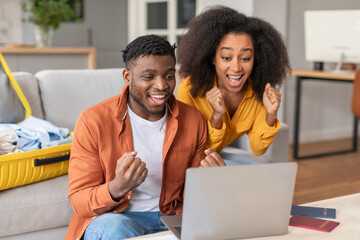 Joyful black travelers couple shaking fists looking at laptop indoor