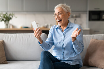 Emotional mature woman looking at cellphone joyfully at living room