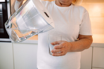 Smiling senior woman pouring filtered water into glass, kitchen interior. Attractive older woman in casual outfit holding glass of fresh water, copy space. High quality photo