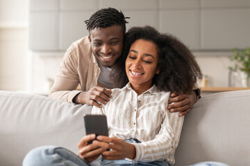 Cheerful black couple sharing social media on cellphone at home