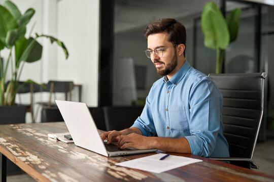 Busy professional business man executive, young male corporate worker, latin businessman wearing glasses typing on computer technology using laptop searching on web working at office workplace desk.
