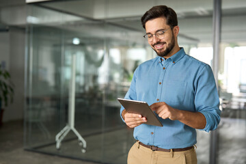 Smiling busy young latin business man manager using tablet computer, happy hispanic businessman executive looking at tab device analyzing finance trading market data working standing in office.