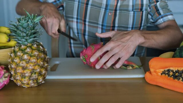 Young man sitting at table surrounded by exotic tropical fruits cutting dragon fruit or pitahaya on a chopping board at home. Healthy lifestyle concept