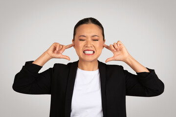 Frustrated Asian businesswoman in a black suit with eyes closed, plugging her ears with fingers