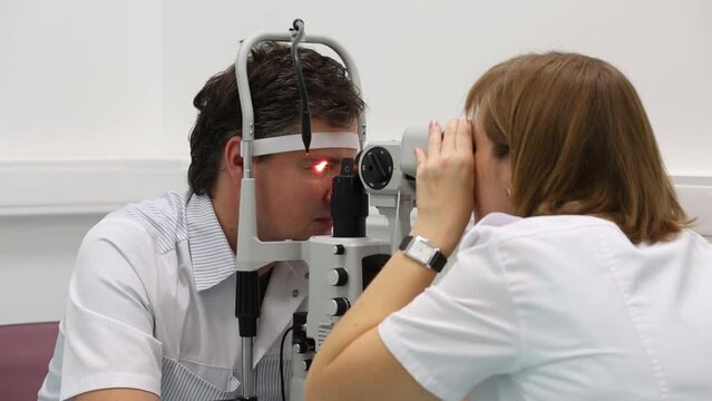Man is sitting near slit lamp and doctor inspecting his eyes.