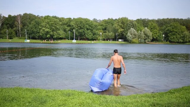 Man in swim briefs go in water of pond with inflatable sofa near lawn