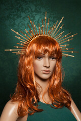 Mannequin with bright long red hair wearing a spiky golden crown posing on a green background