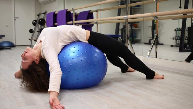 Woman lie and roll on back on rubber blue ball near mirror in gym.