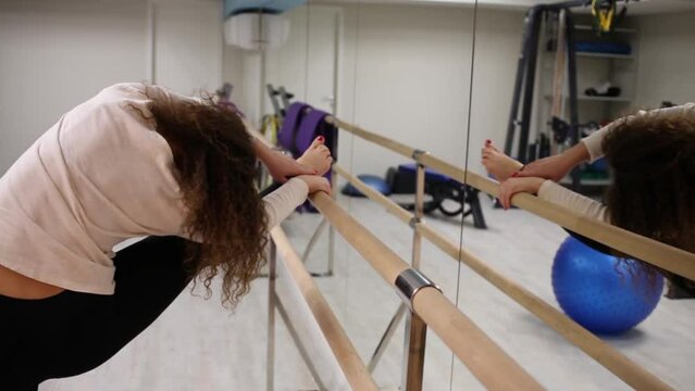 Woman bend to leg on railing near mirror in gym alone.