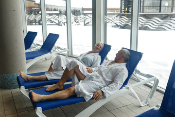 An elderly woman with a man in bathrobes on a sun lounger by the pool.