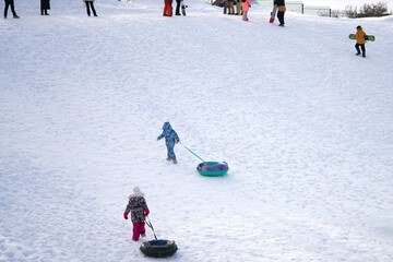 Children skate down a slide on the bank of a frozen pond on inflatable sleds and children's snowboards. Parents are waiting for them at the top of the slope. Copy space.