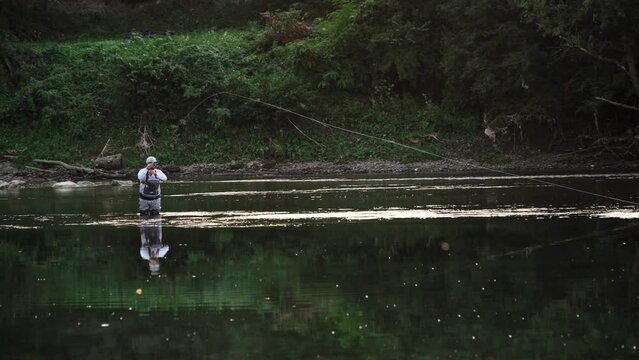 A fisherman in white clothes and cap walks through water with rod catching fish