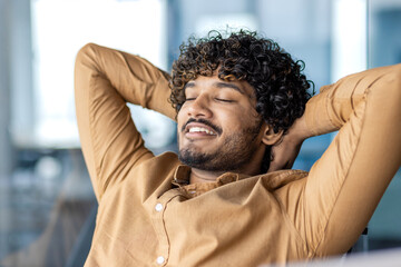 Portrait of relaxed latino guy in camel shirt leaning on chair back and holding hands behind head with wide smile. Productive entrepreneur having break from working on startup with impressive profit. - obrazy, fototapety, plakaty