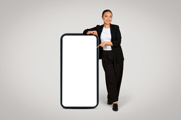 Dynamic Asian businesswoman in professional attire playfully leaning on a giant smartphone with a blank screen