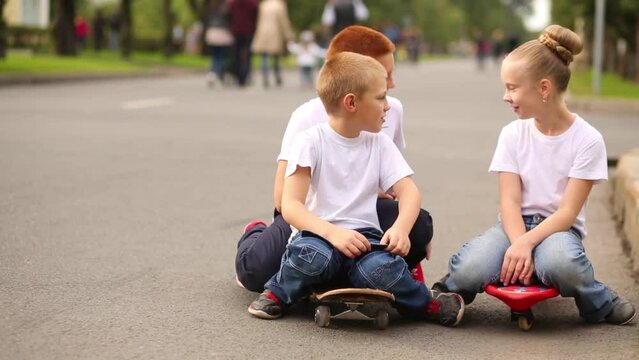Two boys and a girl are sitting on skateboards in the street and talking