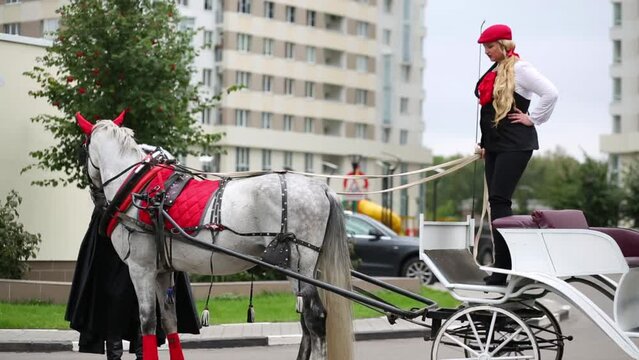 Blond woman and coachman are holding by ribbons a white horses carriage outside.